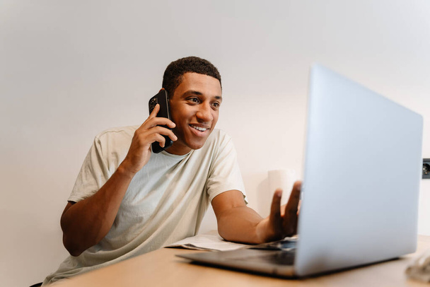 Black man talking on cellphone while working with laptop at desk in hotel - Photo, Image