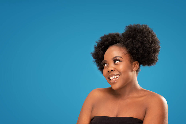 Beauty portrait of african young woman with clean healthy skin looking away, posing on blue studio background. Curly black hair. Real people emotions. A lot of copy space - Zdjęcie, obraz