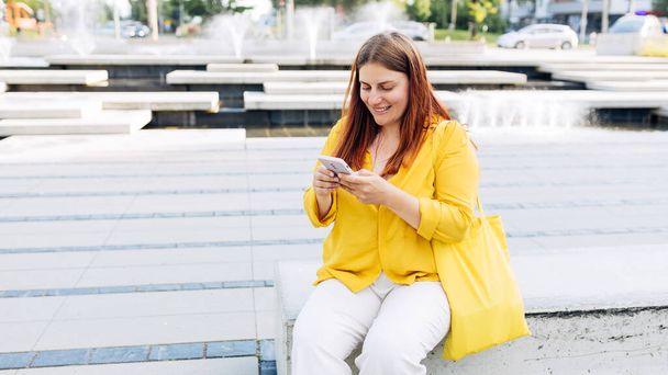 Mooie stijlvolle vrouw zittend op de bank in de stad op de zomerdag en met een smartphone. Telefooncommunicatie. Het concept van stedelijke levensstijl. Reiziger, Controleer sociale netwerken - Foto, afbeelding