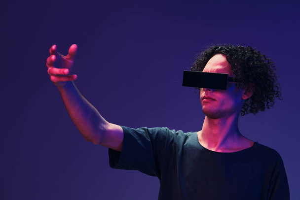 Excited stylish tanned curly man in black t-shirt eyewear touches invisible object posing isolated on color pink blue background. Cool fashion offer. Virtual Reality New Collection concept. Copy space - Photo, Image