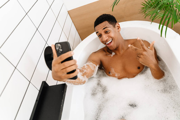 Black man gesturing and using cellphone while taking bath with foam at hotel - Foto, Bild