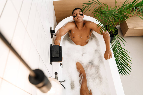 Black naked man wearing eye patches taking bath with foam at hotel - Foto, imagen