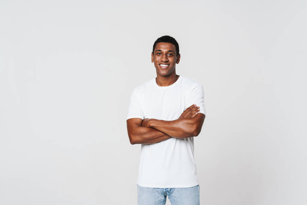 Joven hombre negro sonriendo mientras posando con los brazos cruzados aislados sobre la pared blanca - Foto, imagen