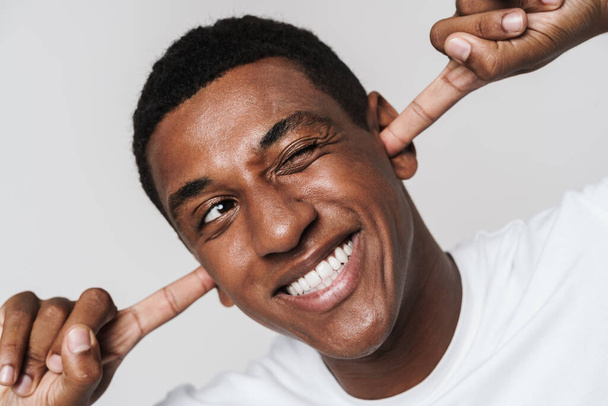 Young black man smiling and winking while plugging his ears isolated over white wall - Valokuva, kuva