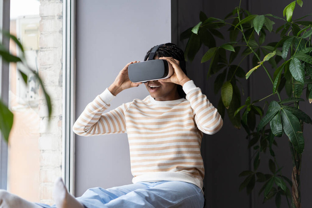 Привіт майбутнє. Молода здивована збуджена чорна жінка, яка розслабляється вдома з віртуальною реальністю, розслаблена африканська жінка використовує занурювальні технології VR, сидячи на підвіконні в затишному будинку з великою кількістю рослин
 - Фото, зображення
