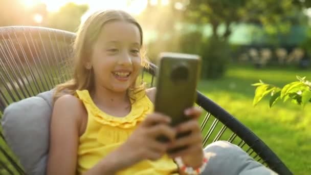 Happy kid girl holding smartphone having video call with friend distantly, online talk in the park outdoor, child using smartphone at home garden, backyard, sunlight, communication, technology concept - Felvétel, videó