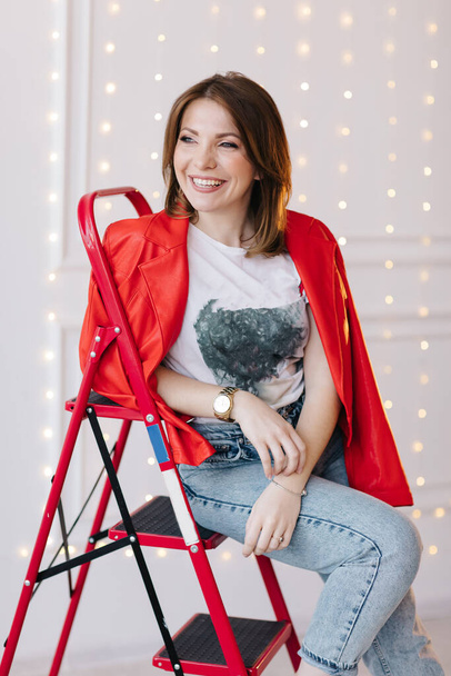 stylish young woman in white t shirt, jeans and red jacket and shoes on high heels is sitting on red ladder. studio short. - Фото, изображение