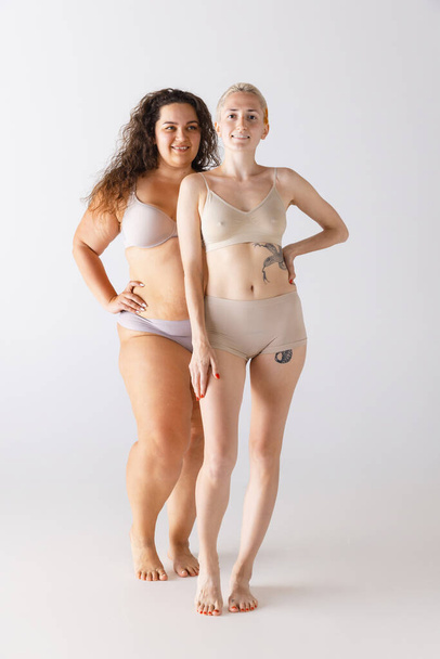 Portrait of two beautiful women, pale and tanned, posing in underwear isolated over grey studio background. Concept of beauty, body and skin care, health, plastic surgery, cosmetics, ad - Fotó, kép