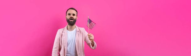 Portrait of a satisfied young man with a beard with an American flag on a red studio background. Great US patriot and defender of freedom. - Photo, Image