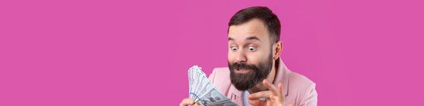 Porträt eines zufriedenen jungen Geschäftsmannes mit Bart und rosa Jacke, der uns Dollarnoten vor rotem Studiohintergrund zeigt. Geschmack, Geruch von Geld. - Foto, Bild