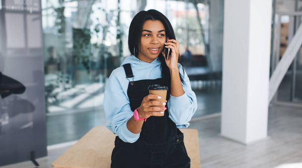 Black girl talking on mobile phone and drinking coffe. Concept of modern woman lifestyle. Young female person with wristwatch standing by wooden table. Blurred interior of modern office - Фото, изображение