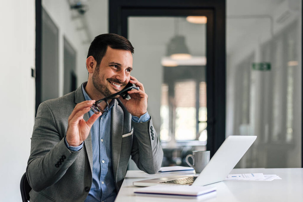 Smiling professional talking on smart phone. Young businessman is working on laptop at desk. He is holding eyeglasses while sitting at desk in office. - Photo, Image