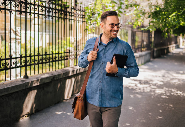 Hombre emprendedor sonriente con tableta digital mirando hacia otro lado. Joven viajero profesional que lleva formales está llevando bolsa de ordenador portátil. Está caminando en la acera de la ciudad.. - Foto, Imagen