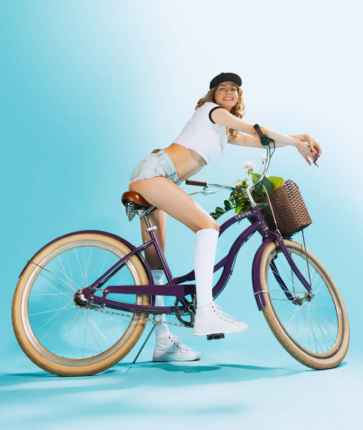 Portrait of beautiful, smiling, young girl sitting on retro bike, posing isolated over blue studio background. Summertime vacation. Concept of summer, fashion, retro style, emotions, freedom, fun - Foto, imagen