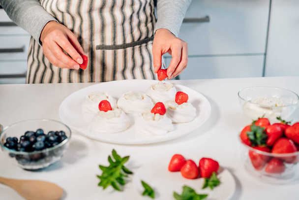 Cooking, making homemade meringue. Female hands decorating pavlova cakes with fresh berries on the kitchen table with ingredients. Recipes for delicious light desserts. Selective focus - Foto, Bild