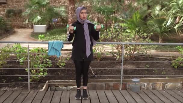 молода красива мусульманка займається спортом з вагами, гантелі на ганку з садом у місті
 - Кадри, відео