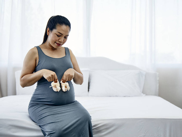 Маленьке взуття для ненародженої дитини з краваткою; вагітна жінка тримає маленьке дитяче взуття, що розслабляється сидить на ліжку в білій спальні
 - Фото, зображення