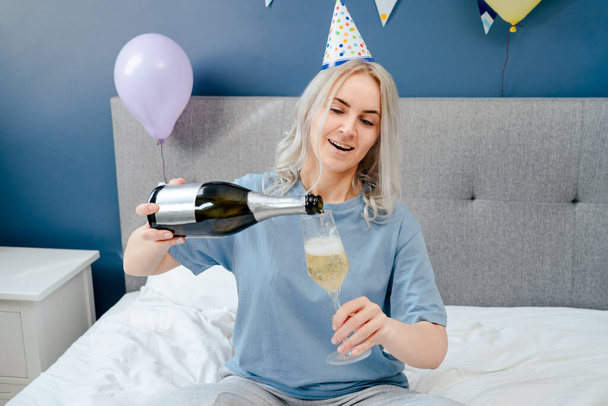 Happy emotional woman in pajama and party cap pouring champagne into glass, sitting on the bed with festive cake in a decorated bedroom. Time for yourself. Celebration at home. Happy birthday concept - Foto, imagen
