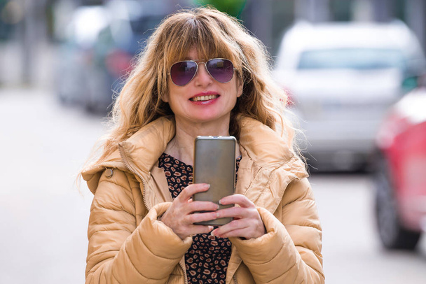 usmívající se žena v slunečních brýlích a kabátu pomocí telefonu na ulici - Fotografie, Obrázek