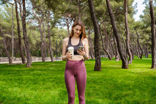 Aranyos kaukázusi nő sportos ruhában a Városligetben, a szabadban egy kávésbögrével a kezében és mobiltelefonnal. Szabadtéri sportok, egészséges életmód. - Fotó, kép