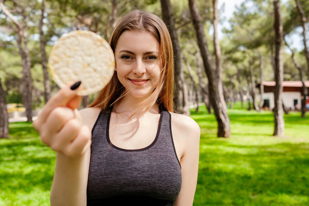 Brünette, sportliche Frau mit schwarzem Sport-BH steht auf dem Stadtpark und schaut in die Kamera, wie sie gesunde Reisknacker isst, lächelt glücklich und positiv. - Foto, Bild