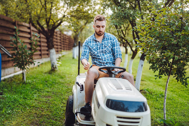 Gardener worker using lawn tractor and cutting grass through garden - Фото, изображение