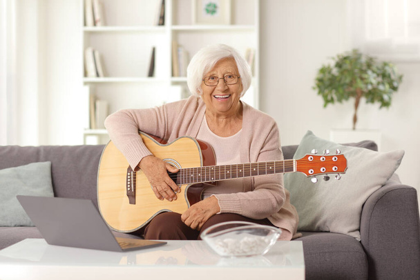 Ältere Frau sitzt zu Hause auf einem Sofa mit einer Akustikgitarre und einem Laptop auf einem Tisch - Foto, Bild