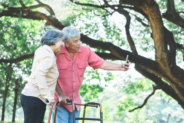 Coppia asiatica di anziani, in piedi con un bastone da sostenere, felici insieme rilassandosi nel giardino verde, E utilizzando il telefono cellulare per scattare una foto, concetto di coppia anziana in vacanza con la natura. - Foto, immagini