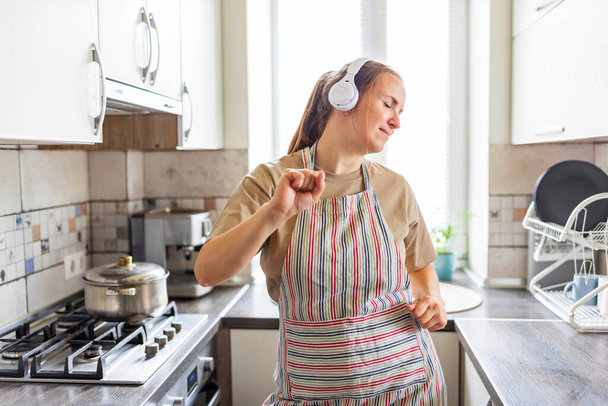 Smiling beautiful woman dancing in kitchen listening music with headphones. - Photo, image