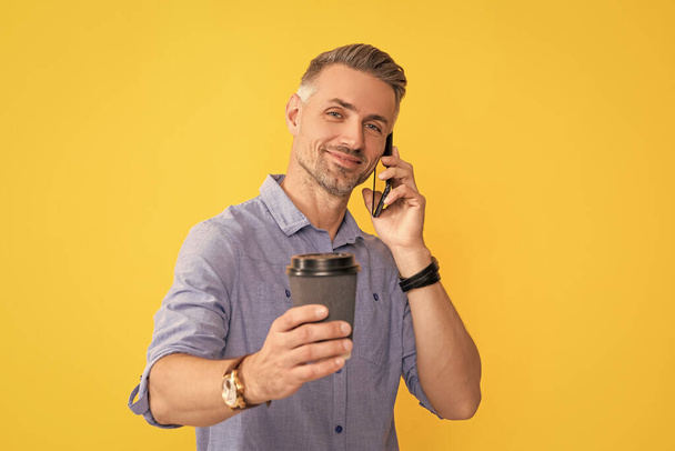 sonriente hombre maduro hablando por teléfono con taza de café sobre fondo amarillo, enfoque selectivo, café de la mañana. - Foto, Imagen