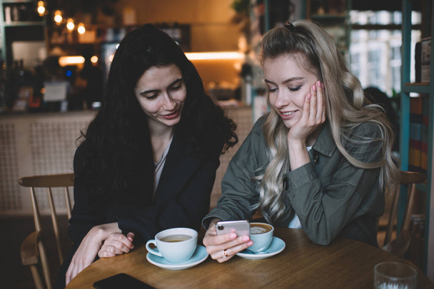 Concentrated multiracial female friends in casual clothes sitting at table with cups of coffee in cafe together and using mobile phone thoughtfully - Valokuva, kuva