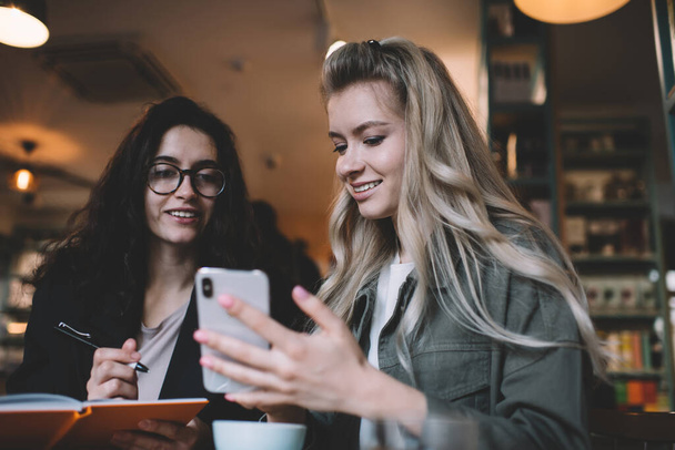 Young smiling women in casual outfit sitting at table in modern workspace and discussing business ideas while looking at screen of mobile phone - Valokuva, kuva