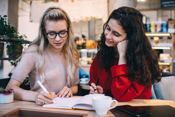 Vrolijke vrouwelijke collega 's in comfortabele kleding met notebook komen met zakelijke strategie voor het opstarten terwijl zitten aan tafel in coffeeshop - Foto, afbeelding