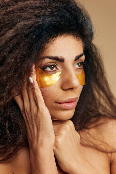 Die attraktive Latina mit goldenen Hydrogelpflastern unter einer Gesichtsmaske mit Augenkontakt schaut zur Seite und posiert isoliert auf pastellbeigem Hintergrund. Kosmetikprodukt ad Natural beauty Skincare concept - Foto, Bild