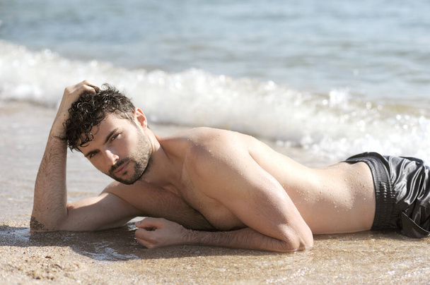 Young sexy man portrait lying near ocean, topless body, man relax on a sand - Photo, image