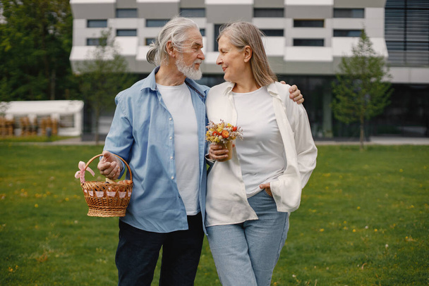 Caucasian elderly couple standing on a grass in summer. Man holding straw basket and woman flowers. Woman wearing white shirt and man blue one. - Foto, immagini