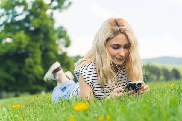 Vaalea valkoihoinen tyttö lepää nurmikolla ja tarkistaa sosiaaliset verkostot. Täysi laukaus ulkona. Laadukas kuva - Valokuva, kuva