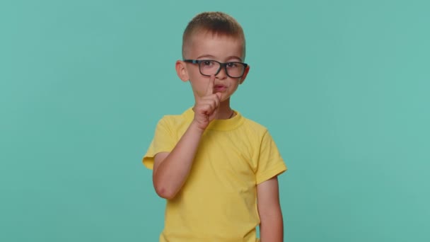 Stil zijn, alsjeblieft. Kleine peuter kinderen jongen drukt index vinger tot lippen maakt stilte gebaar teken vertelt geen geheim. jong kleuter in bril geïsoleerd alleen op blauw studio achtergrond - Video