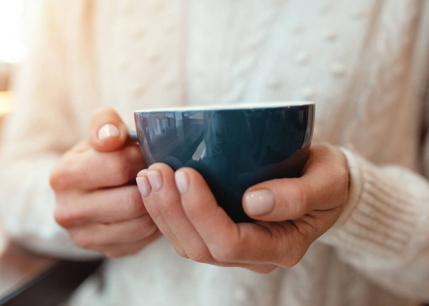 Retrato de una mujer joven en sweter blanco con una taza de café en la mano mientras que en la cafetería Frío manos gusano café - Foto, imagen
