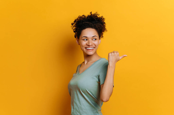Joyful pretty african american curly haired young woman in basic t-shirt, looks and points finger to the side behind the back, at empty copy space, stands on isolated orange color background, smiling - Foto, afbeelding