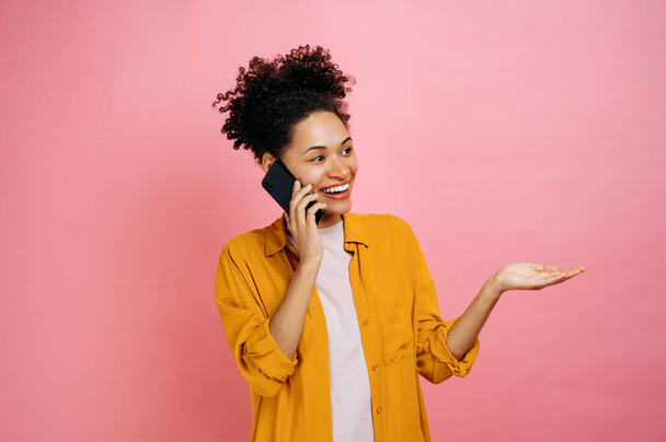 Τηλεφωνική ομιλία μέσω κινητού. Χαρούμενη χαρούμενη αφρικάνικη νεαρή γυναίκα, έχει μια ευχάριστη τηλεφωνική συνομιλία, χαρούμενη έκφραση προσώπου, στέκεται σε απομονωμένο ροζ φόντο, χειρονομώντας, χαμογελώντας. - Φωτογραφία, εικόνα