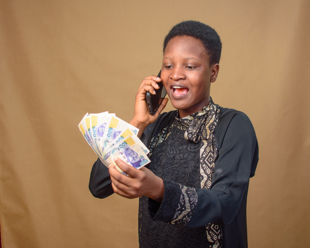 African Nigerian lady excited as she stares at the spread of multiple naira notes, cash, currency or money in her hands while making a call with a smart phone - Foto, Bild