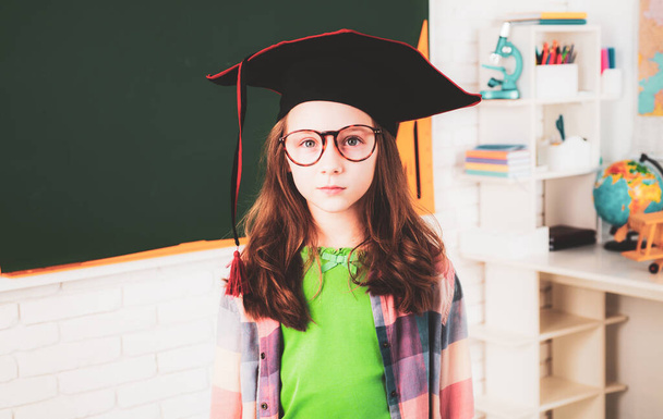 Education and school concept - little student girl studying at school. Portrait of cute schoolgirl with graduation hat in classroom. Graduate college, preschool or university cap - Photo, Image
