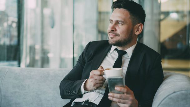 Smiling bearded Caucasian businessman in formal clothes smiling, relaxing and looking at his smartphone screen while drinking coffee in airy cafe during lunch time - Фото, изображение