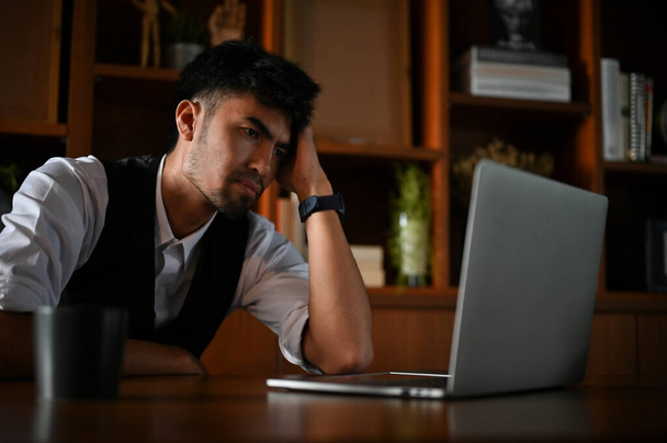 Müde und gelangweilt sitzt ein asiatischer Geschäftsmann oder Manager an seinem Schreibtisch vor dem Laptop. - Foto, Bild