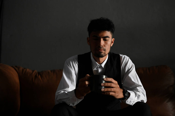 Профессиональный многолетний азиатский бизнесмен или мужчина исполнительный директор в официальном костюме сидит на темном кожаном диване со своим кофе. - Фото, изображение
