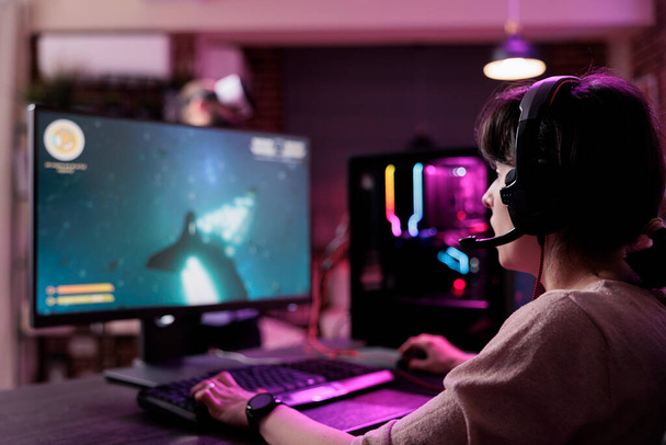 Modern insan oturma odasında bilgisayar ve neon ışıkları kullanarak çevrimiçi video oyunları oynuyor. Kadın oyuncu sanal atış turnuvasında aksiyon oyunuyla eğleniyor. - Fotoğraf, Görsel