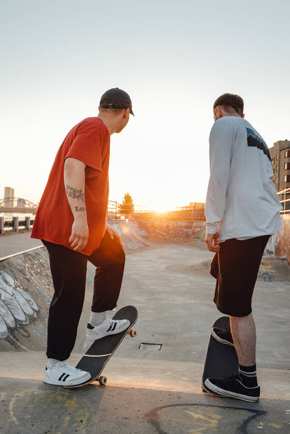 Aufnahme von zwei tätowierten Hipster-Jungen beim Skateboarden im Skatepark bei Sonnenuntergang. - Foto, Bild