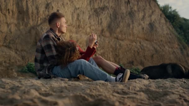 Nuori pari dating auringonlaskun meren rannalla. Nätit rakastavaiset katsomassa kuvia älypuhelimella. Rento nainen ja mies käyttävät matkapuhelinta ulkona. Hipsterit nauttivat kesälomasta yhdessä. Elämäntapakäsite - Materiaali, video