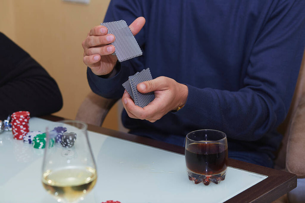 Τα χέρια του ανθρώπου ανακατεύουν τα χαρτιά σε ένα παιχνίδι πόκερ. Πατατάκια, κάρτες, ένα ποτήρι ουίσκι στο τραπέζι με αντανάκλαση. Λέσχη πόκερ. - Φωτογραφία, εικόνα